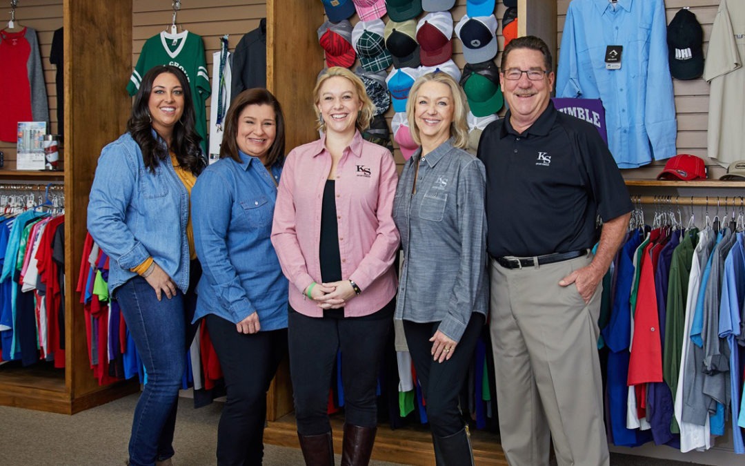 The team of K&S Sportswear standing in front of custom screen printing shirts, embroidered hats, and promotional products.